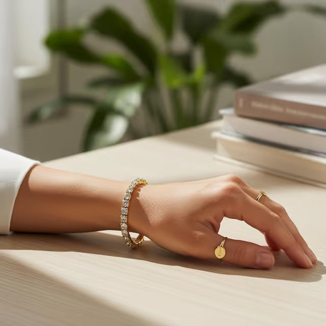 Hand wearing a gold bracelet and ring on a light surface with books and a plant in the background