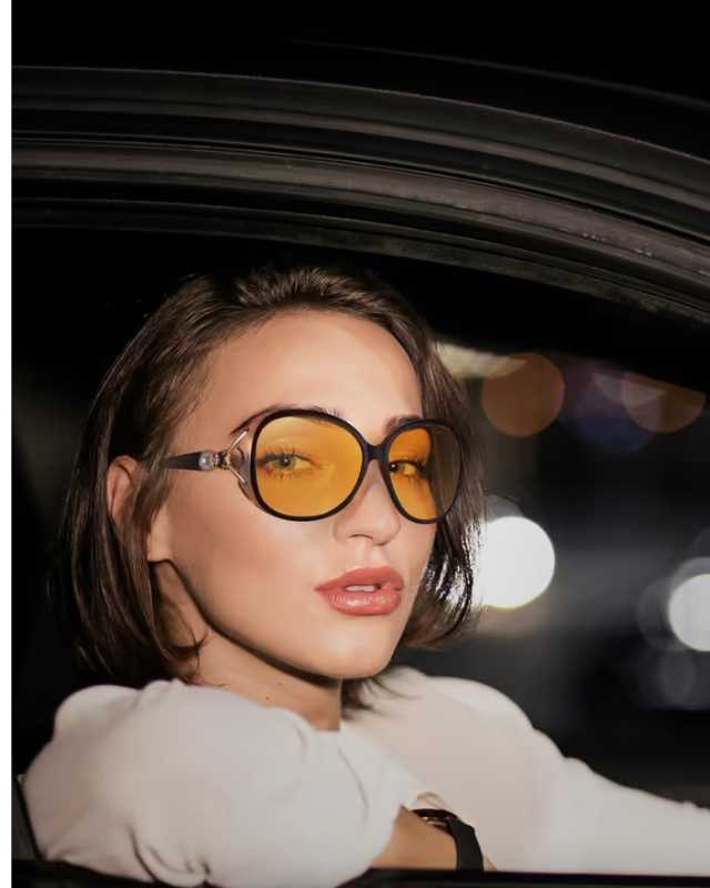 Woman wearing yellow sunglasses inside a car with blurred lights in the background