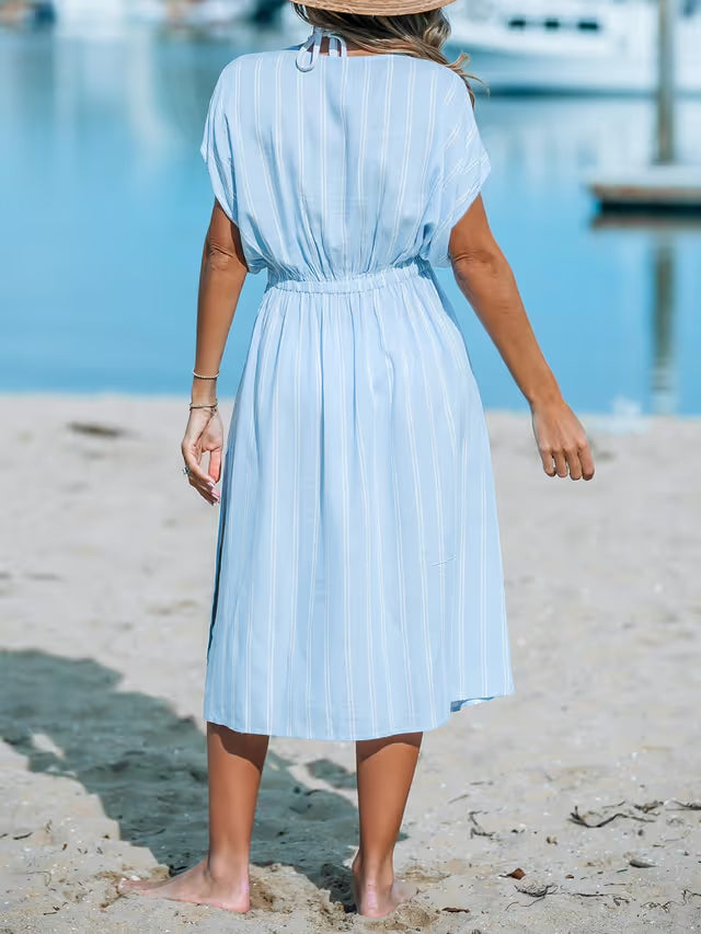 Woman in a light blue dress standing on a sandy beach with water and boats in the background.